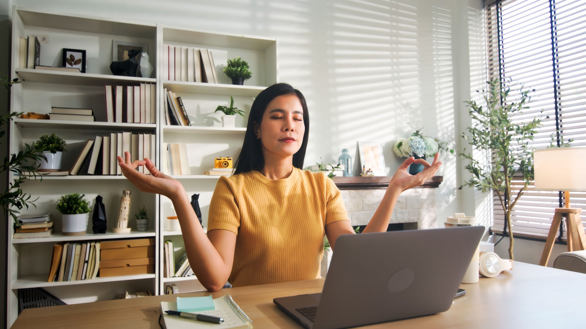 A young woman takes a break from work to meditate at her home office desk. She sits with eyes closed, hands in a meditative pose, surrounded by books and plants in a serene environment.